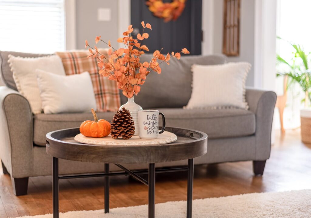 Living room decorated in warm autumn colors, with a coffee table centerpiece tray holding a pinecone, small pumpkin, mug, and a vase filled with orange leafy branches, creating a cozy and frugal fall decoration.